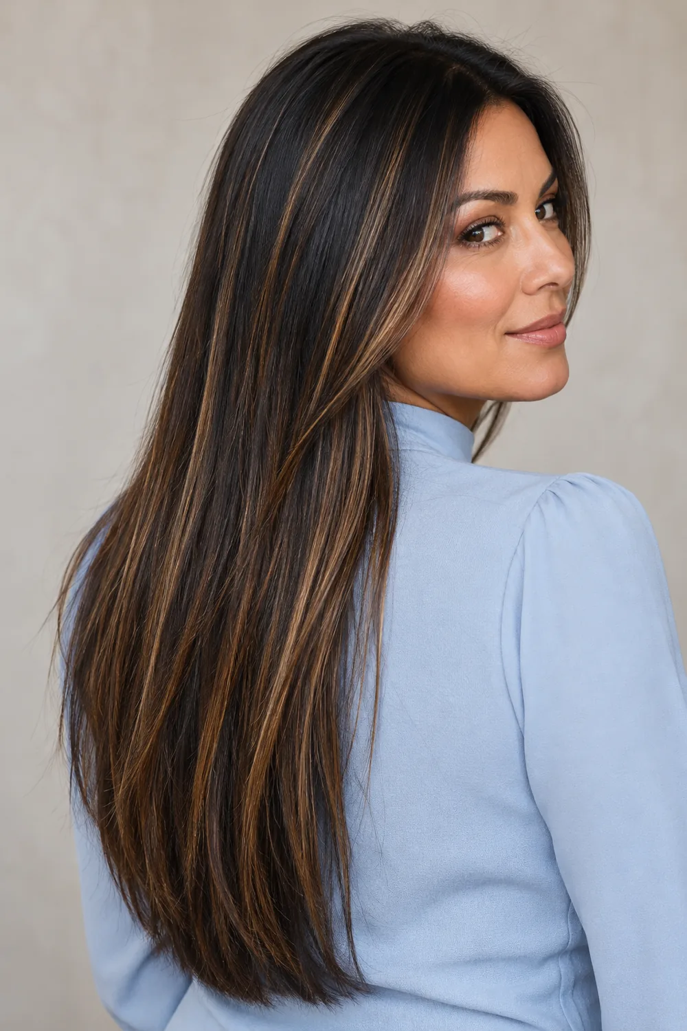 A woman with long, straight black hair threaded with fine caramel highlights, seen in three-quarter view against a neutral background.