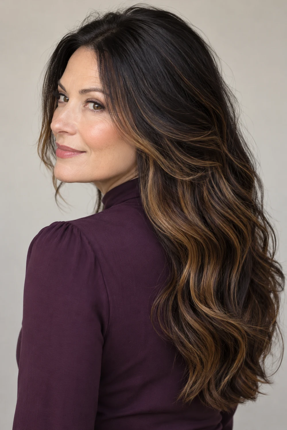 A woman in a plum high-neck blouse looks over her shoulder, showing long black waves with caramel ribbons through the side and lower lengths.