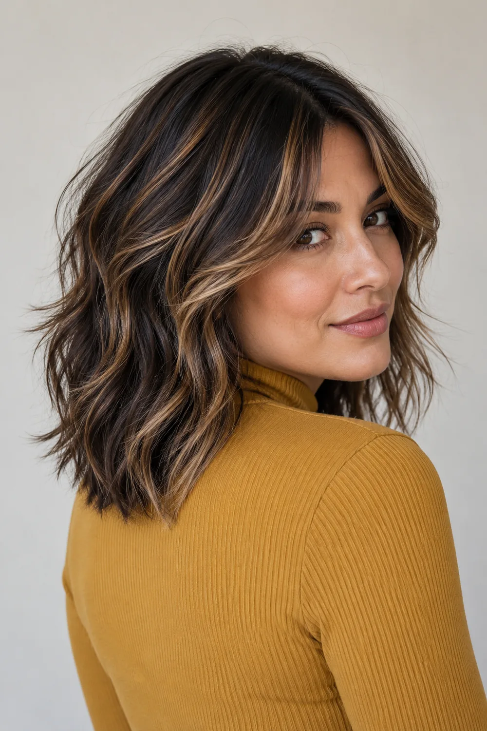Adult woman in a rear three-quarter portrait, wearing a mustard high-neck top, with a shoulder-length dark shag and caramel highlights through the fringe and layers.