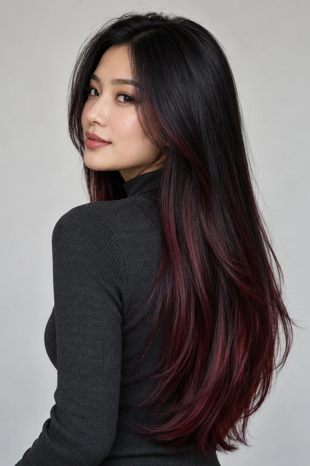An adult woman in a charcoal high-neck top looks over her shoulder, showing long, straight black hair with burgundy highlights through the lengths.