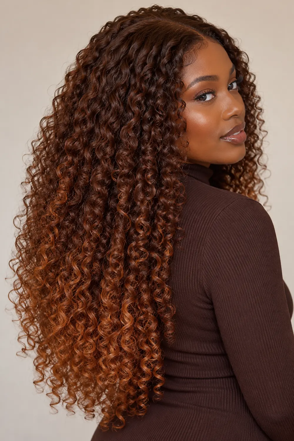 A woman with deep brown skin in a three-quarter portrait, wearing long defined chestnut brown curls that gradually brighten to copper-gold through the lengths.