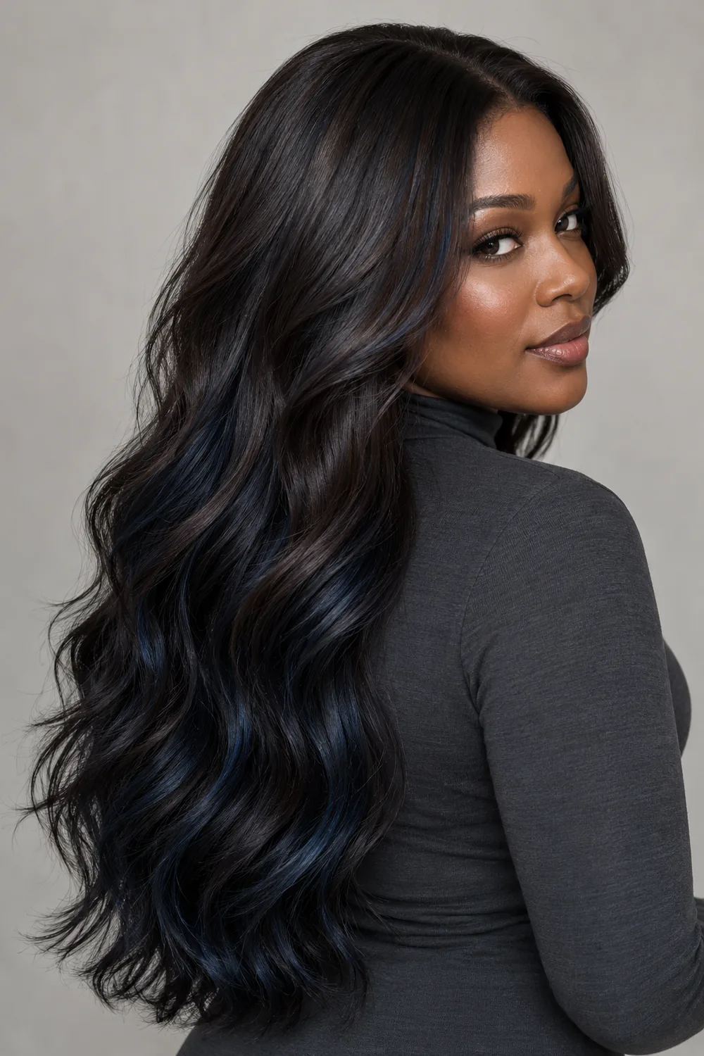 A woman with a deep complexion looks over her shoulder, showing long, loose inky espresso waves with subtle blue-black ribbons and a charcoal high-neck top.