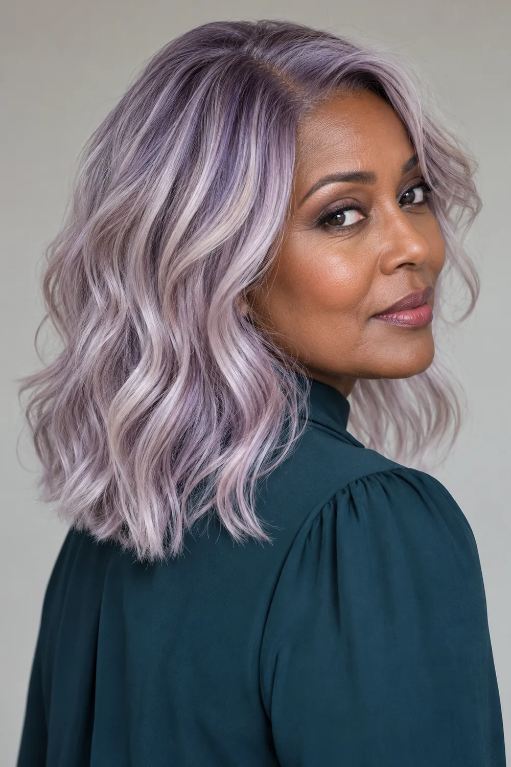 A woman with medium-deep cool skin wears a softly waved lavender-blonde long bob with smoky lilac roots and silver-violet ribbons, styled with a dark teal high-neck blouse.