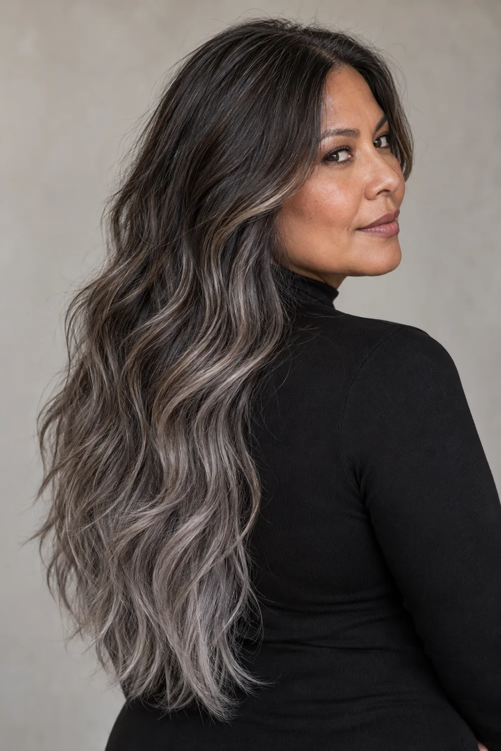 A woman in a black high-neck top looks over her shoulder, showing long wavy hair that fades from charcoal-brown roots to smoky pewter ends with ash highlights.