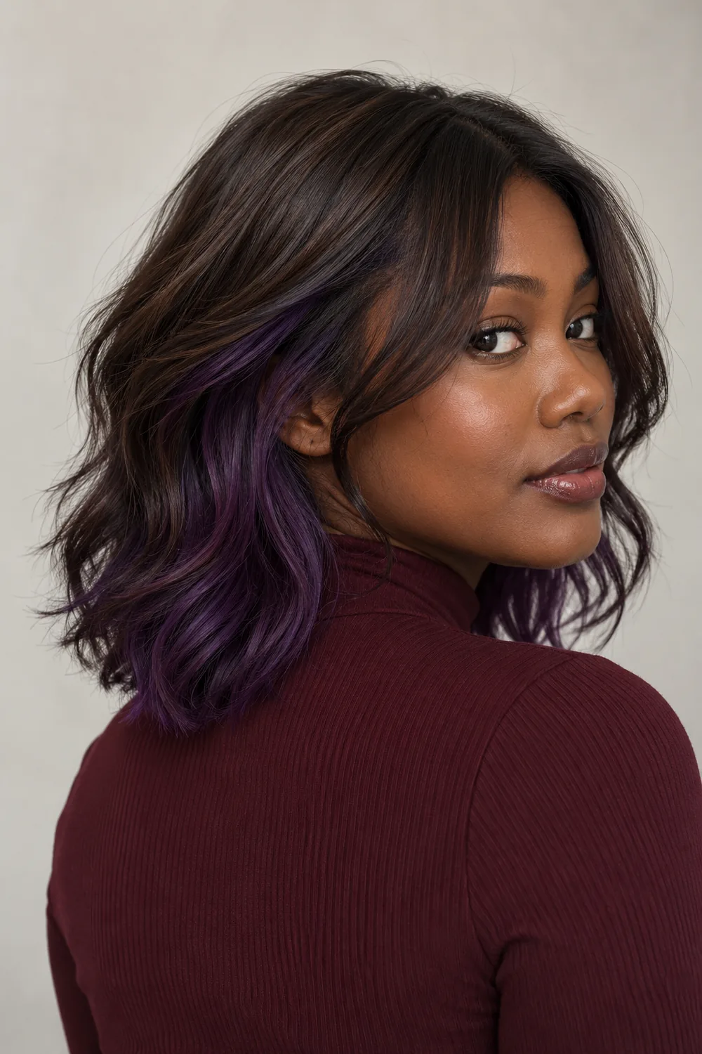 Adult woman in a rear three-quarter portrait with a dark brown wavy shag and a visible deep violet underlayer, wearing a burgundy high-neck top.