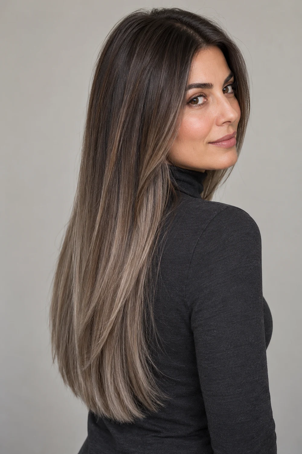 A woman with light-medium olive skin looks over her shoulder, showing long, straight cool brunette hair with muted mushroom taupe balayage and smoky ends.