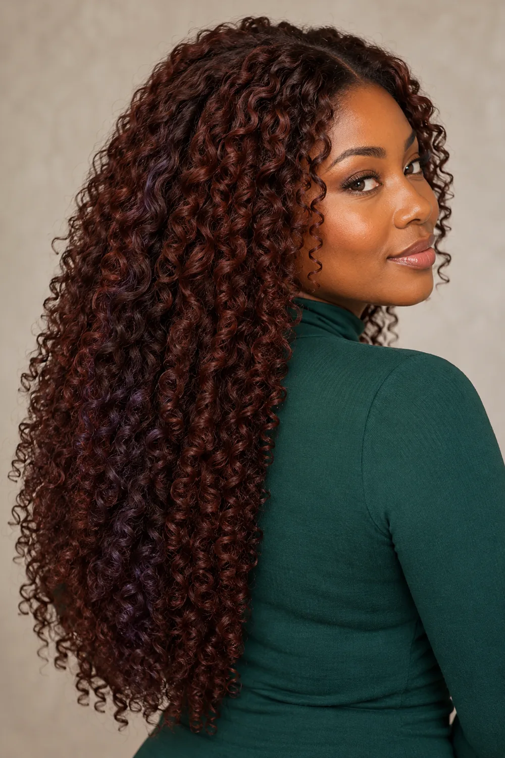 A woman with deep olive-brown skin looks over her shoulder, showing long defined mahogany curls with subtle violet-brown lowlights and an emerald high-neck top.