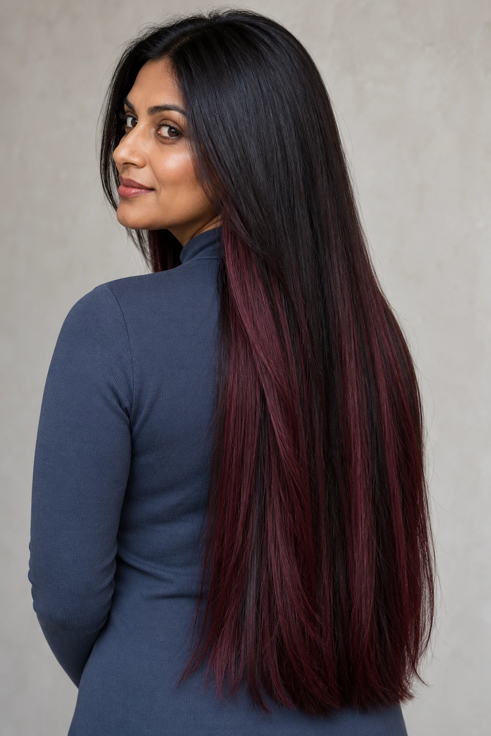 A fictional adult Indian woman with deep olive skin looks over her shoulder, showing long sleek near-black hair with black-cherry red-violet panels through the lower lengths and ends.
