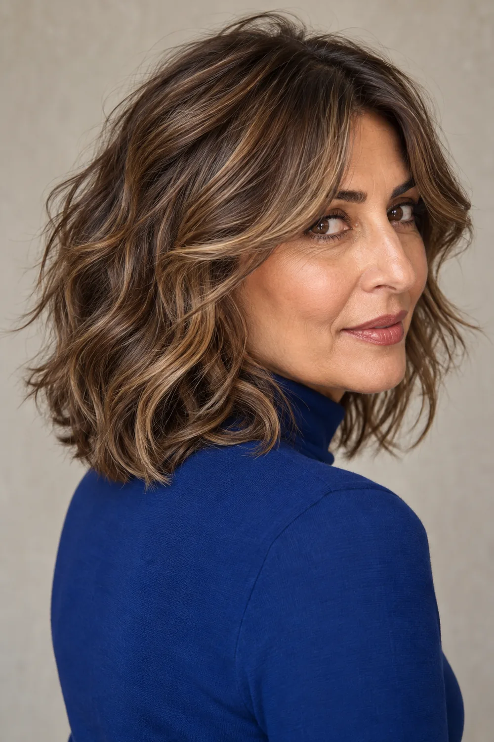A fictional adult woman with medium olive skin wears a shoulder-length wavy shag with curtain fringe, medium brown roots, and softly blended dark blonde ribbons. She looks over her shoulder in a cobalt high-neck top against a neutral background.