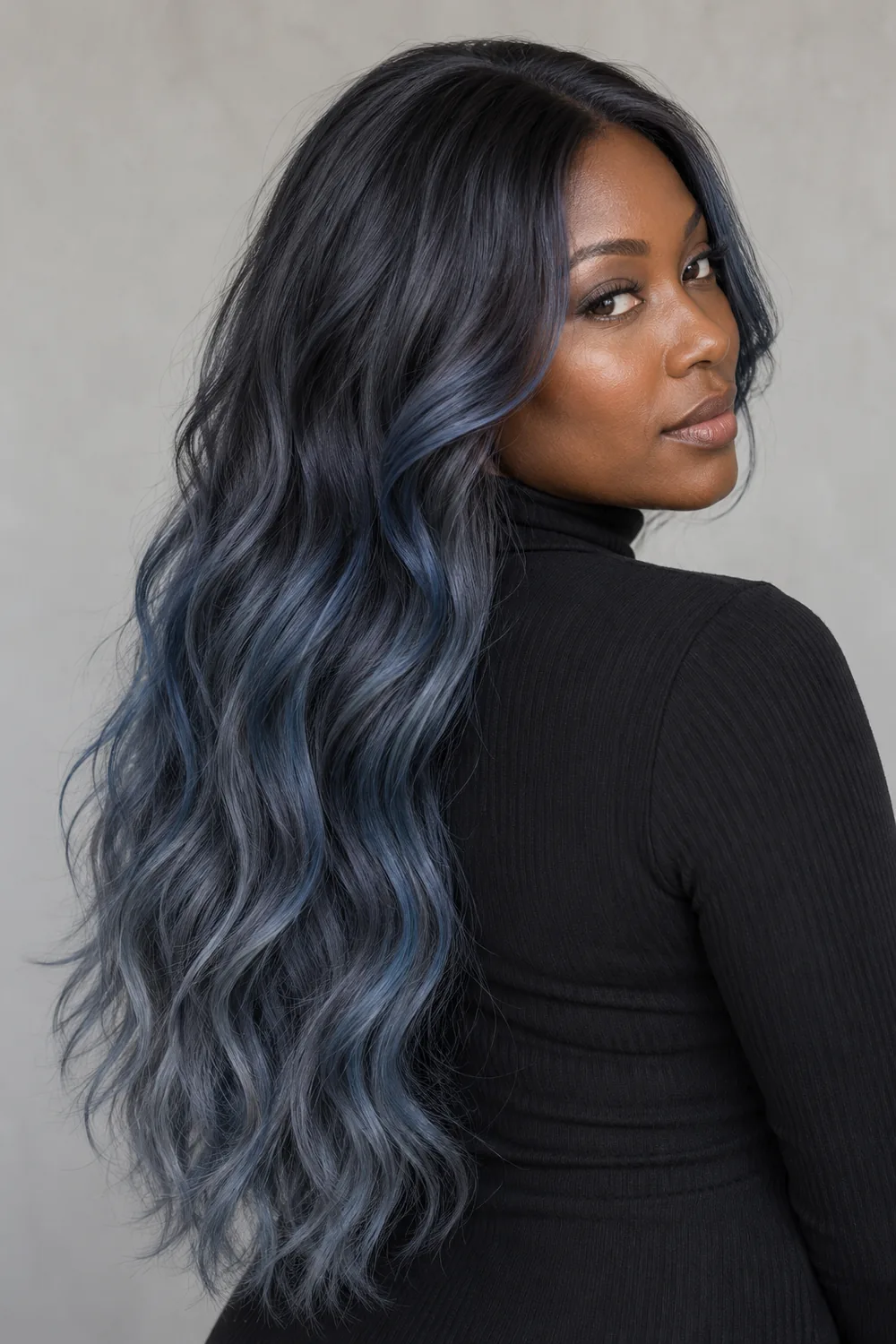 A Black woman in a black high-neck top, shown in three-quarter view with long loose waves transitioning from charcoal roots to muted smoky blue-gray ends.