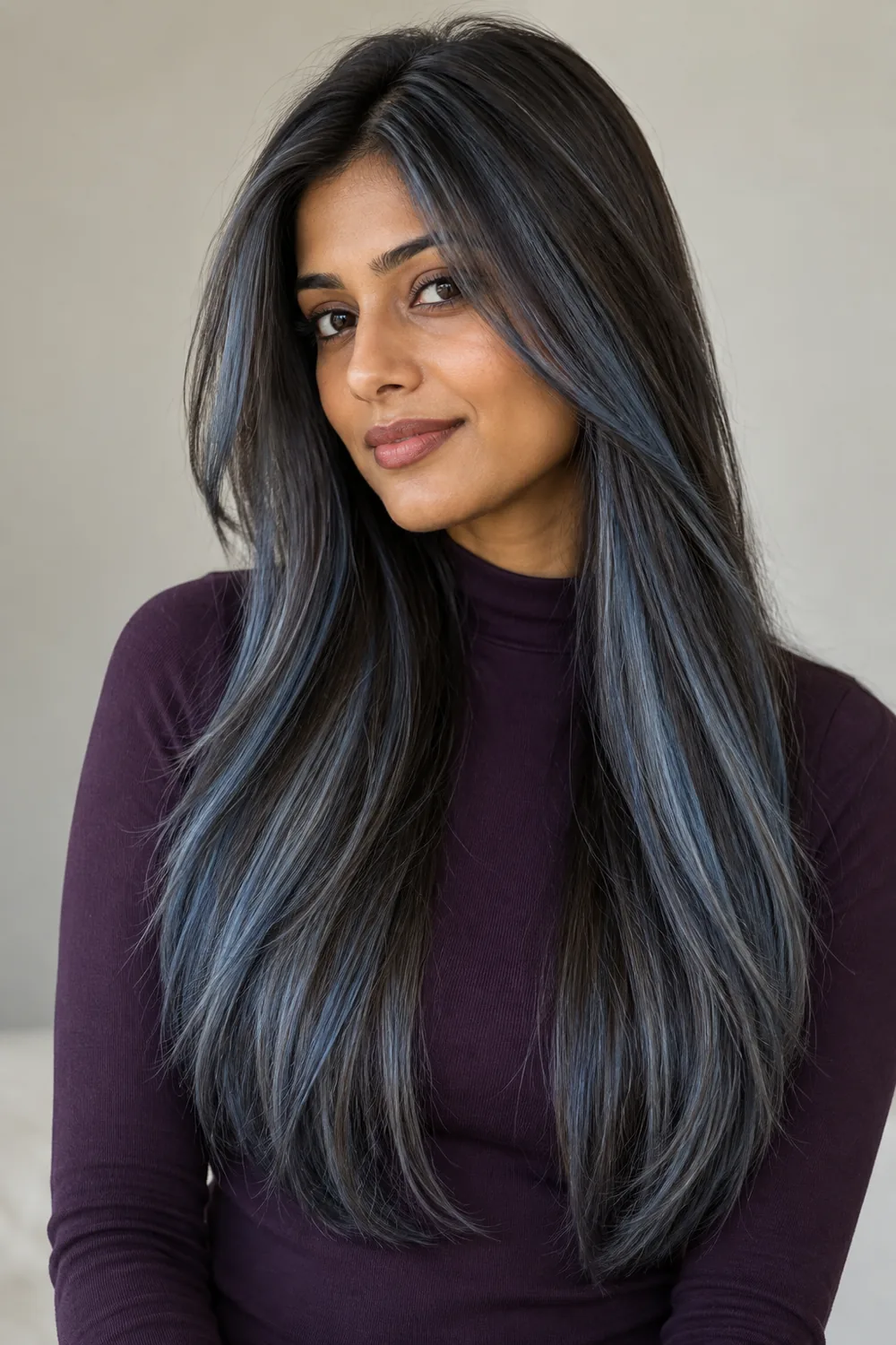 An adult South Asian woman in a plum high-neck top wears long, straight dark hair with fine smoky blue-gray face-framing ribbons and scattered cool strands through the lengths.