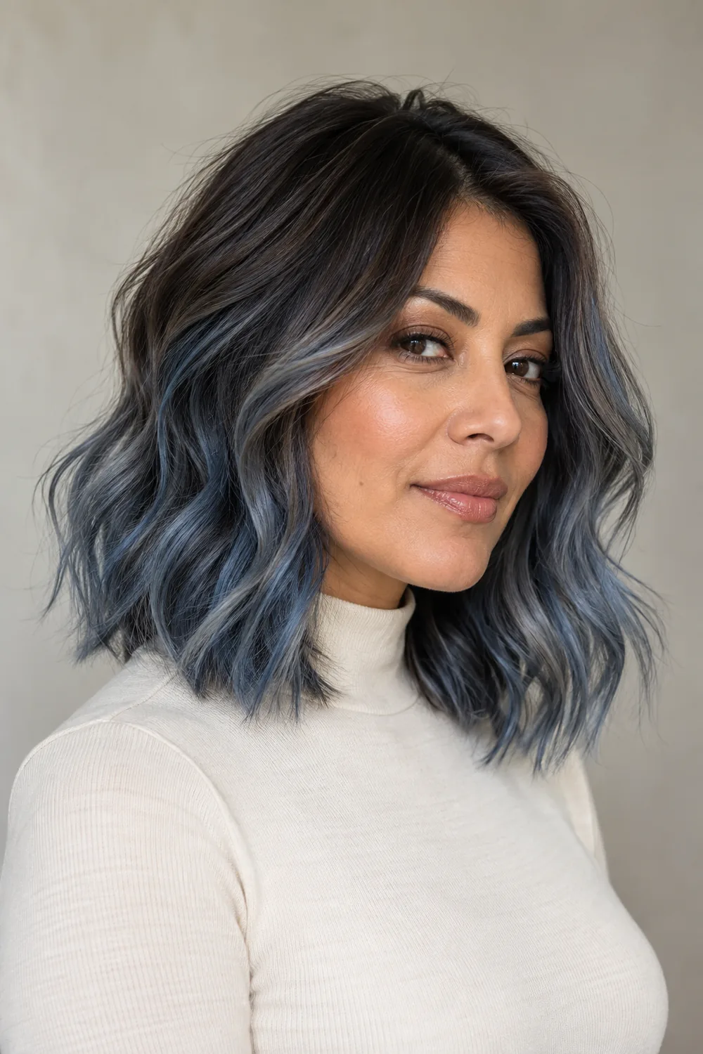 Adult woman with olive-brown skin wearing a cream high-neck top, shown in a three-quarter portrait with a wavy shoulder-length lob, espresso roots and smoky slate-blue balayage.