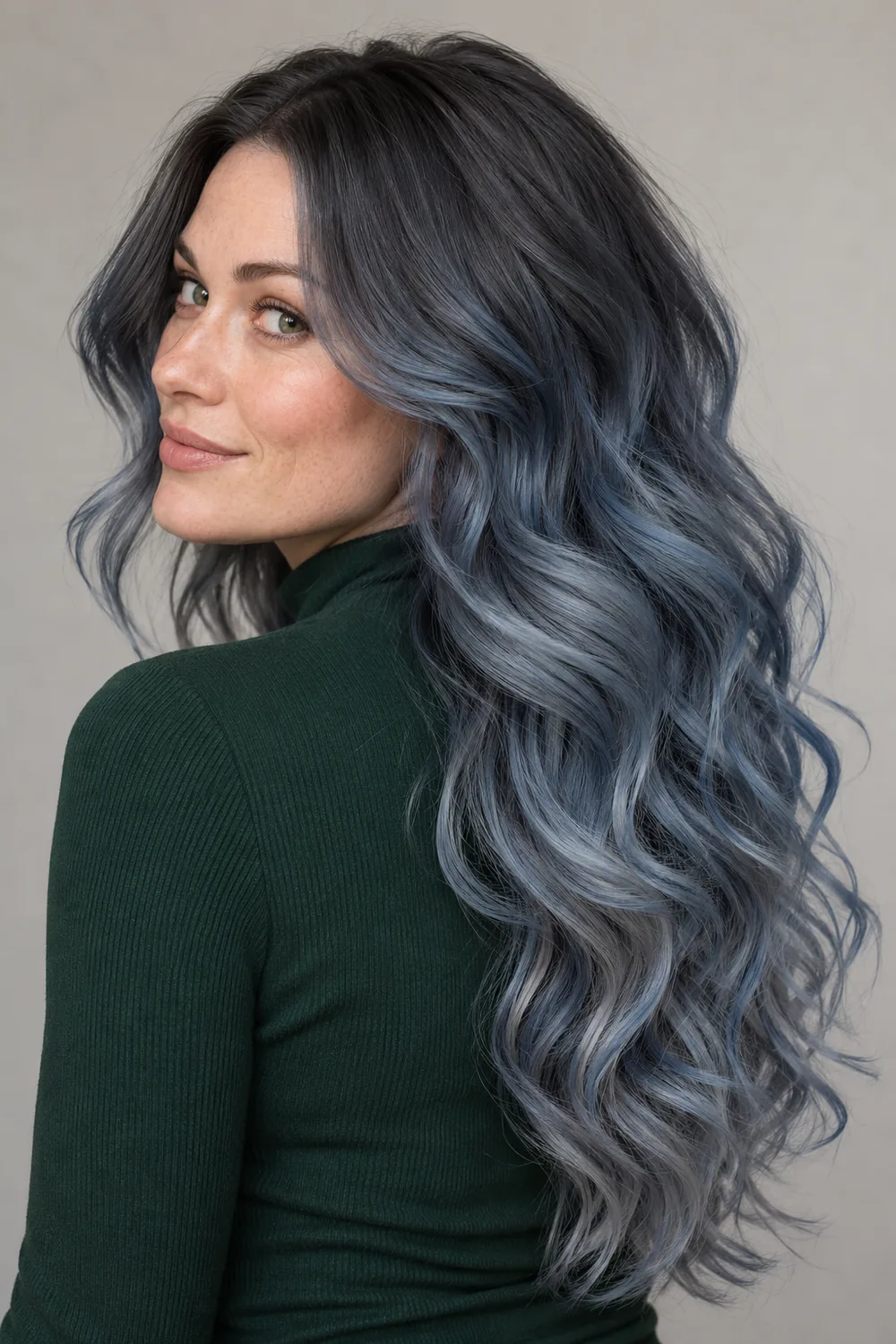 A freckled adult woman in a forest-green high-neck top, with long loose curls transitioning from dark graphite roots to smoky steel-blue gray ends.