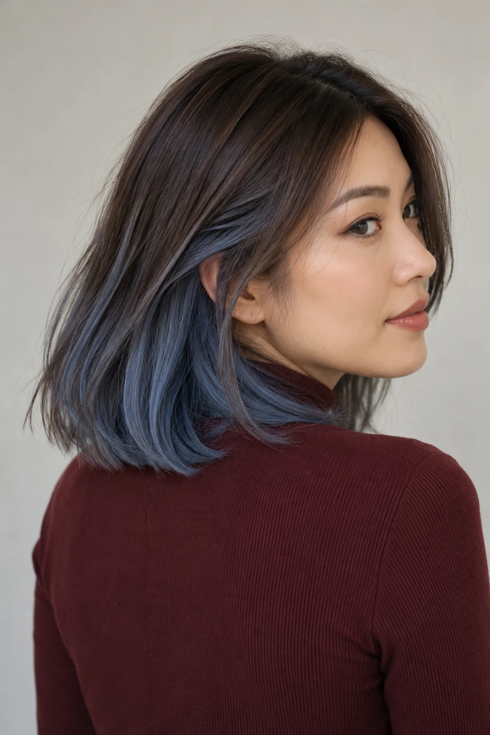An adult East Asian woman in a burgundy high-neck top, turning to show shoulder-length dark ash-brown hair with smoky slate-blue underlayers.