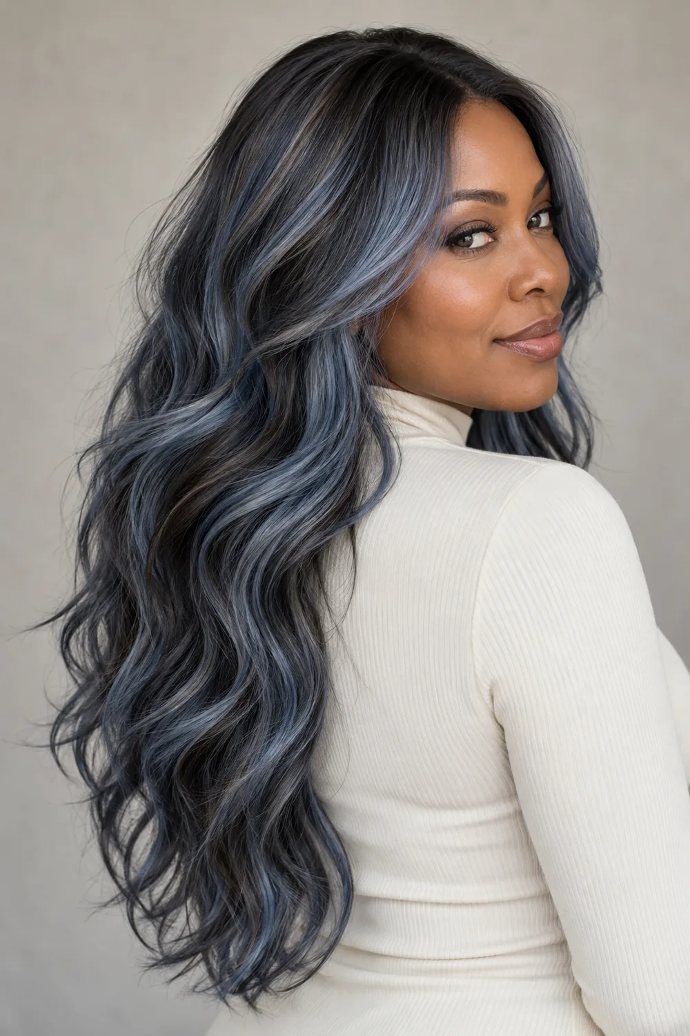 A Black woman in an ivory high-neck top looks over her shoulder, showing long smoky blue-gray waves with charcoal lowlights and dark blended roots.