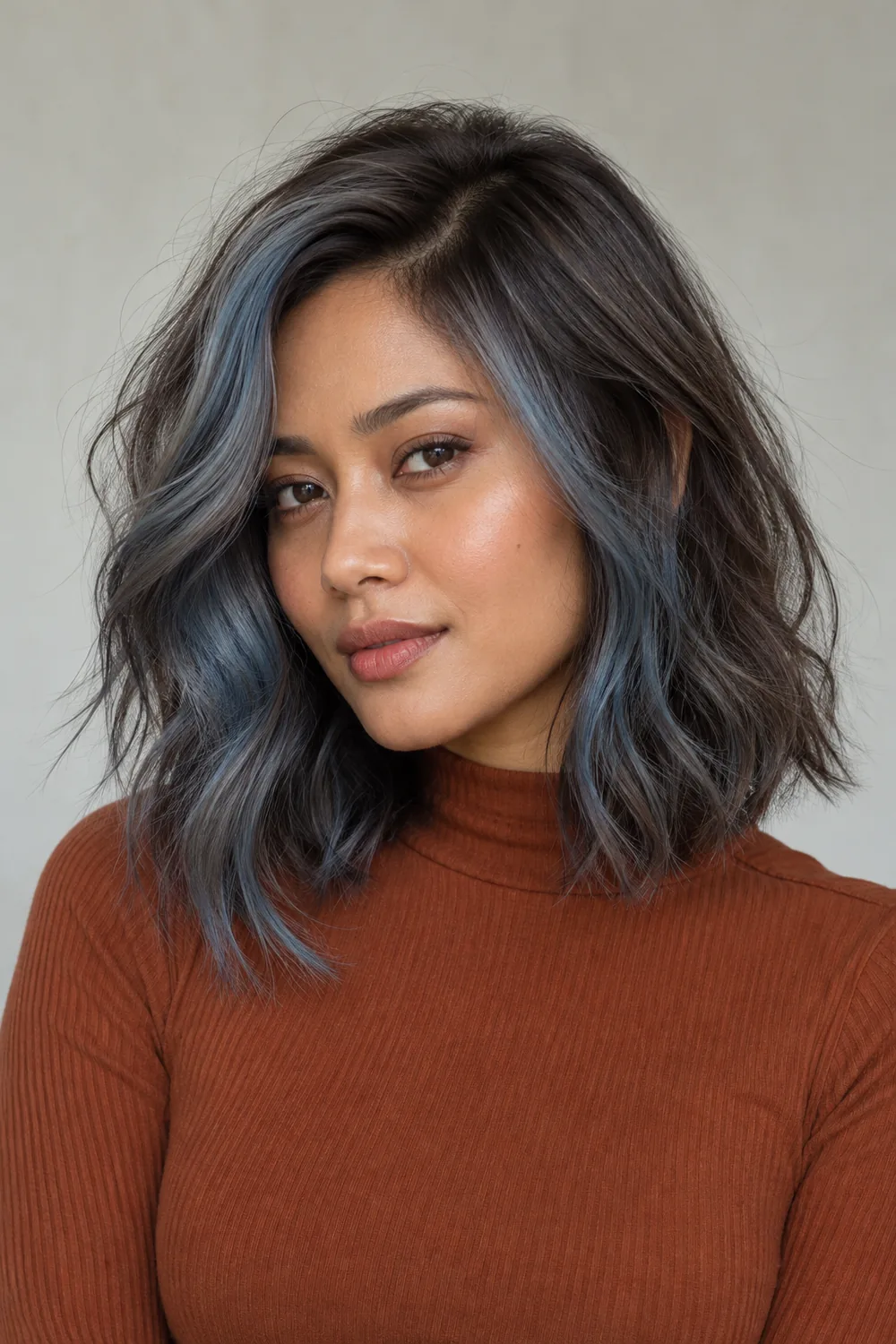Adult woman with a collarbone-length wavy ash-brown shag and asymmetrical smoky blue-gray face-framing sections, wearing a rust-colored high-neck top.