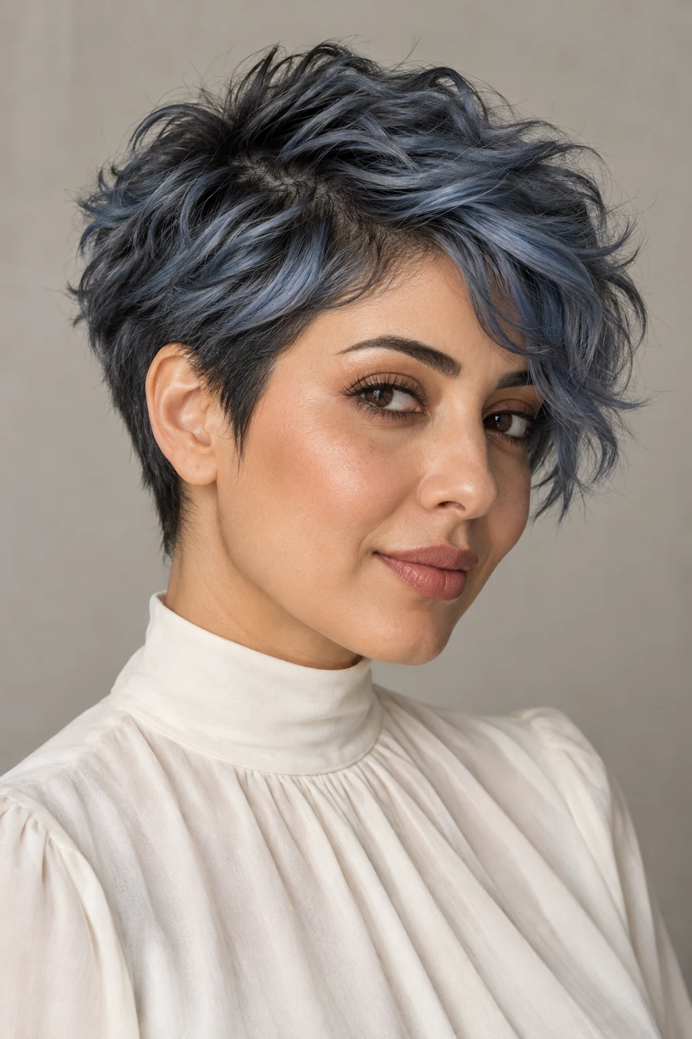 Adult woman in a cream high-neck blouse with a textured pixie, smoky blue-gray top and fringe, and darker charcoal roots and tapered sides.