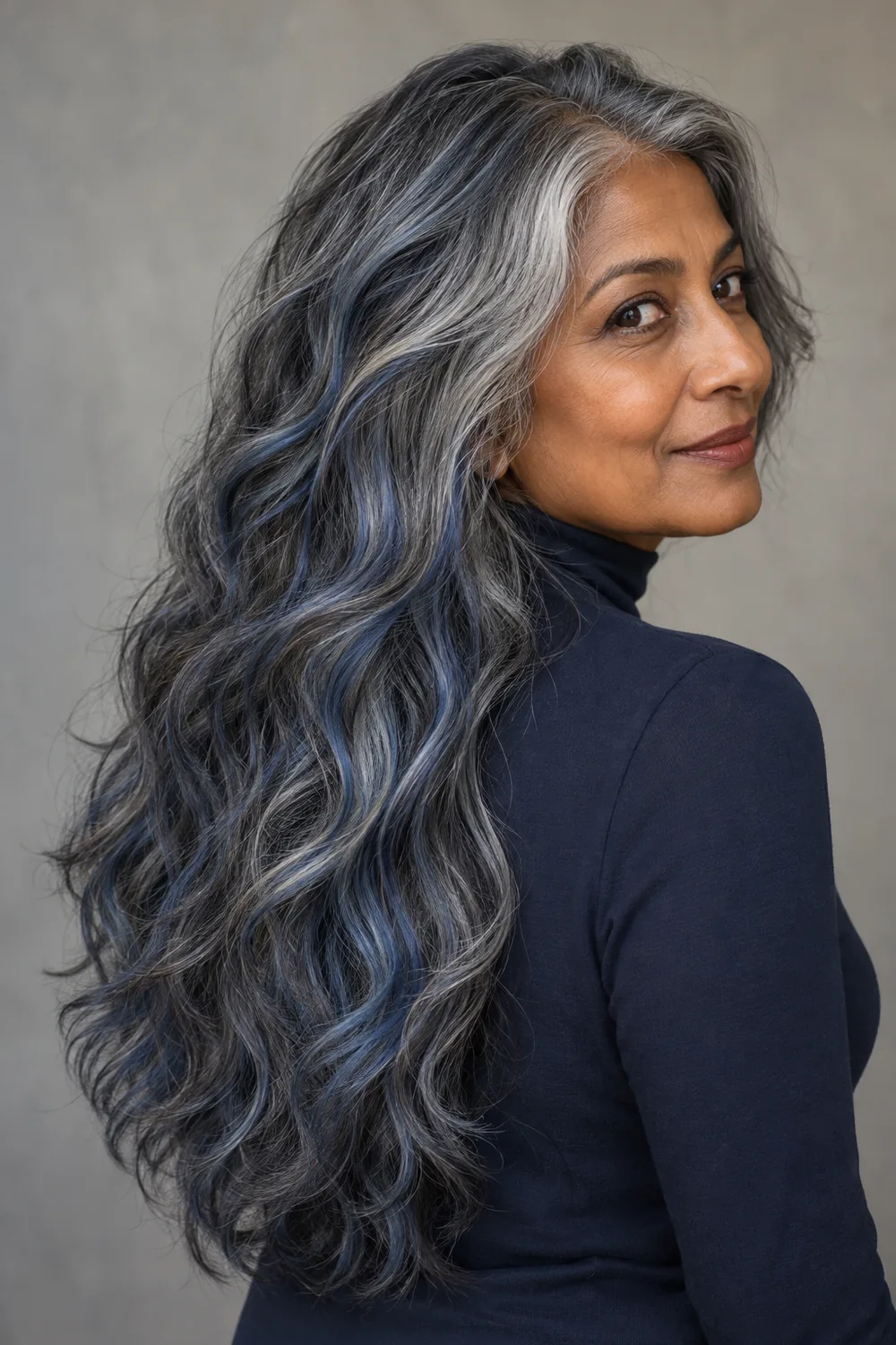 A mature South Asian woman in a navy high-neck top, turning over her shoulder to show long wavy salt-and-pepper hair with smoky blue-gray ribbons.
