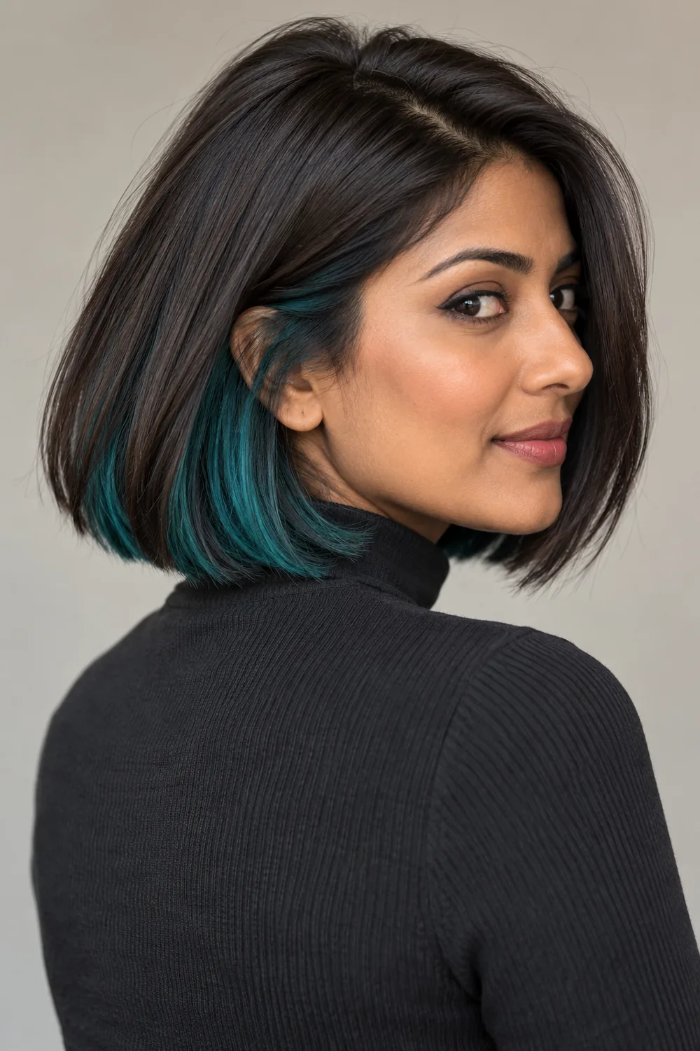 A woman in a charcoal high-neck top looks over her shoulder, revealing teal underlayers beneath her dark espresso bob.