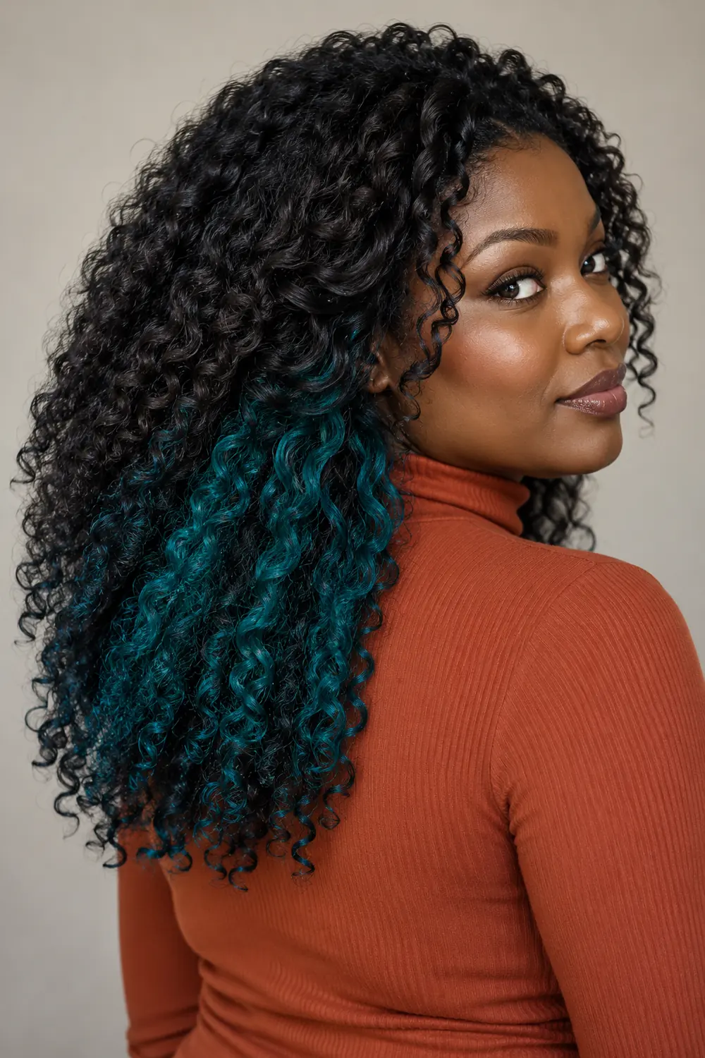 A woman with long black coily curls and peacock teal peekaboo sections beneath the lower layers, wearing a rust high-neck top.
