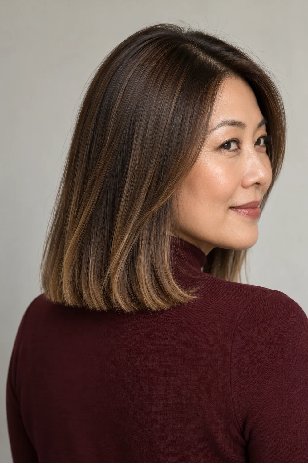 Adult East Asian woman in a burgundy high-neck top, shown in a three-quarter rear view with a blunt shoulder-length teddy bear brunette bob and softly golden-beige ends.